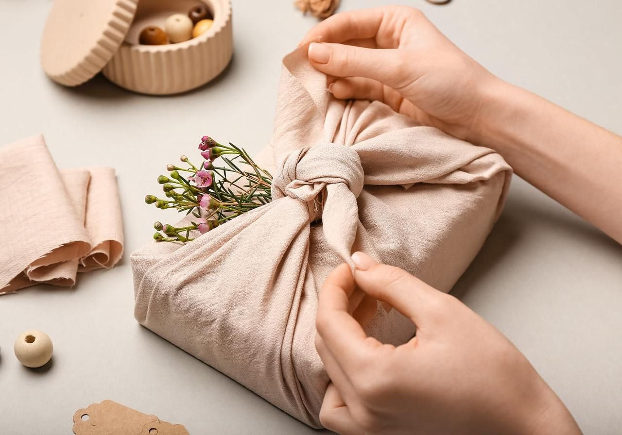 Hands tying a furoshiki fabric wrap around a gift, embellished with small pink flowers.